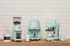 Three Smeg coffee appliances are arranged on a wooden shelf against a white brick wall. A coffee grinder, a drip coffee maker, and an espresso machine are displayed. Náhled obrázku produktu