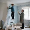 A father and son are wallpapering a room. The son is on a ladder applying wallpaper, while the father holds a measuring tape. Produktbild-Vorschau