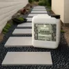A pathway made of stepping stones and dark gravel, with a large white container of Roxlin Botanic Kiesbinder in the foreground. Produktbild-Vorschau