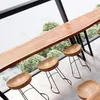 A long wooden table with several wooden stools in front of a large window with greenery outside. Produktbild-Vorschau