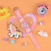 A pink unicorn-themed watch is displayed on an orange background, surrounded by small decorative items. Produktbild-Vorschau