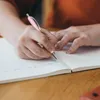 A person's hands are writing in a notebook with a pink pen. Voir les images du produit