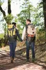 A couple holding hands and walking on a dirt path in a forest, both wearing backpacks. Podgląd zdjęcia produktu