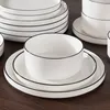 A stack of white ceramic plates and bowls with black trim on a dark brown wooden surface. Voir les images du produit