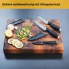 A set of three kitchen knives with black handles and protective sheaths are arranged on a wooden cutting board with chopped herbs, lemon slices, and a garlic bulb. Produktbild-Vorschau