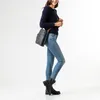 A woman wearing a dark blue sweater and light blue jeans poses with a blue handbag. Produktbild-Vorschau