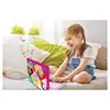 A young girl is sitting on a bed, smiling and interacting with a pink toy laptop. The laptop has images of Disney princesses on it, and musical notes are depicted floating around it. Produktbild-Vorschau