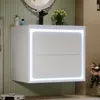 A white wall-mounted bathroom vanity with two drawers, illuminated by LED lighting around its edges. It is set against a light green striped wallpaper. A black faucet, a round mirror with an ornate frame, a vase of flowers, and a soap dispenser are visible Produktbild-Vorschau