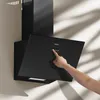 A black kitchen range hood mounted on a wall, with a hand interacting with its control panel. Shadows from foliage are cast on the wall. Voir les images du produit