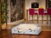 A golden retriever dog is resting on a dog bed in a modern living room with a kitchen island and bar stools in the background. Náhľad obrázku produktu