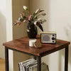 A square side table with a wooden top and black metal legs, featuring a vase of flowers, a candle, and a retro radio. Produktbild-Vorschau