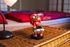 A Knuckles figurine wearing glasses and a smartwatch sits on a woven surface next to a lamp, with a bed in the background. Podgląd zdjęcia produktu