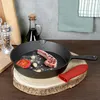 A cast iron pan with a lamb chop, tomato, and garlic is placed on a wooden stand. A bottle of oil and a pepper grinder are in the background. Produktbild-Vorschau