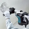 A person wearing white gloves is using a Makita hammer drill to break a hole in a concrete wall. Produktbild-Vorschau