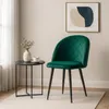 A green velvet chair with black metal legs is placed next to a small black side table with a vase and a cup on it. A framed abstract artwork hangs on the wall behind the chair. Produktbild-Vorschau