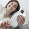 A woman is lying in bed, smiling and holding a clear phone case with a Samsung Galaxy A12 phone inside. Produktbild-Vorschau