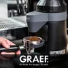 A person's hand holding a portafilter under a coffee grinder. Podgląd zdjęcia produktu