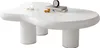 A white, irregularly shaped coffee table with two cylindrical legs, decorated with two candles, a stack of books, and two small decorative spheres. Voir les images du produit