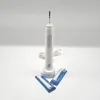 A white electric toothbrush with its charging base and two replacement brush heads in packaging, set against a plain white background. Produktbild-Vorschau