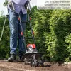 A person is using a tiller in a garden setting with a hedge in the background. Produktbild-Vorschau