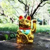 A golden Maneki Neko cat figurine sits on a rock in a natural outdoor setting with trees and greenery in the background. Produktbild-Vorschau