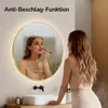 A woman is applying makeup in front of a lit, round bathroom mirror. The mirror has condensation on it, indicating it has an anti-fog function. The bathroom has a modern sink and faucet. Produktbild-Vorschau