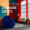 A living room scene with a large navy blue beanbag chair in the foreground and colorful walls and furniture in the background. Produktbild-Vorschau
