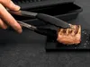 A hand using tongs to place a grilled piece of meat on a dark surface next to a grill. Produktbild-Vorschau