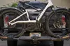 A white electric bicycle is mounted on a black bike rack attached to the back of a dark-colored vehicle. A license plate is visible on the bike rack. Produktbild-Vorschau