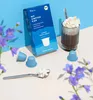 A box of coffee capsules and a glass of coffee with whipped cream, with flowers in the foreground. Produktbild-Vorschau