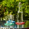 A glass bottle of water and two glasses sit on a table outdoors, with greenery in the background. Produktbild-Vorschau