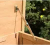 A close-up view of a metal hinge mechanism attached to a wooden structure, with greenery and yellow flowers in the background. Voir les images du produit