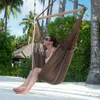 A woman relaxing in a brown hammock chair on a sandy beach with palm trees and a hut in the background. Produktbild-Vorschau