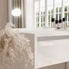 A close-up view of a white, high-gloss desk with a drawer and decorative dried grass in the foreground. The background shows a window with natural light and curtains. Produktbild-Vorschau