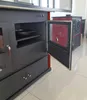 A close-up view of a dark gray wood-burning stove with its oven door open, revealing a red interior and a baking pan on a shelf. Another similar stove is visible in the background. Náhled obrázku produktu