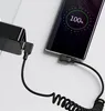 A smartphone is charging from a wall adapter using a coiled USB-C cable. The phone screen shows 100% battery. Anteprima dell’immagine prodotto