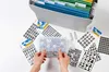 A top-down view of various sheets of stickers with numbers and letters, a clear plastic container with number stickers, and a filing box filled with hanging folders. Produktbild-Vorschau