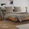 A neatly made bed with taupe bedding and pillows, set in a bedroom with a plant and artwork. Produktbild-Vorschau