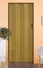 A closed folding door with a wood-like finish, standing in a room with a plant on a shelf to the left and a white chair to the right. Produktbild-Vorschau