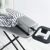 A close-up shot of a gray ironing board with a striped garment draped over it. The ironing board has a small sleeve board attached and a black accessory tray extending from the side. The background is a plain white wall and a light gray floor. Voir les images du produit