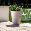 A large beige planter with purple and green flowers sits on a paved patio next to a stone wall. A white table and chairs are visible in the background. Anteprima dell’immagine prodotto