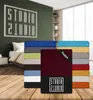 A stack of folded bed sheets in various colors, with one sheet prominently displayed in bordeaux color, placed on a wooden table in front of a blurred bedroom background. Produktbild-Vorschau