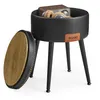 A black storage ottoman with a wooden lid is shown with headphones and a small radio inside. The ottoman is supported by three black legs and has a tan leather logo tag on the front. Náhled obrázku produktu