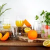 A still life arrangement of stemless glasses, some filled with orange juice and others with ice and fruit, surrounded by fresh oranges and lemons on wooden stands. Produktbild-Vorschau
