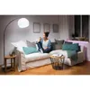 A woman is sitting on a couch in a living room, holding a smartphone and looking at it. The room is decorated with framed pictures on the wall and a floor lamp. Náhled obrázku produktu