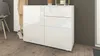 A white, high-gloss chest of drawers with three doors and one drawer, placed against a dark gray wall on a wooden floor. A chair and some shoes are visible to the left. Produktbild-Vorschau