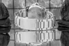 A close-up, black and white shot of the back of a metal watch band and clasp, with a reflection below. Two dark, textured rocks are visible on either side of the watch. Náhľad obrázku produktu