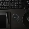 A top-down view of a black AirPods case with a Mercedes logo on a dark, textured surface, next to a keyboard and a stylus on a tablet. Voir les images du produit