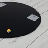 A black circular mirror with mounting hardware attached, lying on a light gray wooden floor. Produktbild-Vorschau