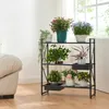 A black metal plant stand with multiple shelves filled with various potted plants, placed indoors near a window and a sofa. Produktbild-Vorschau